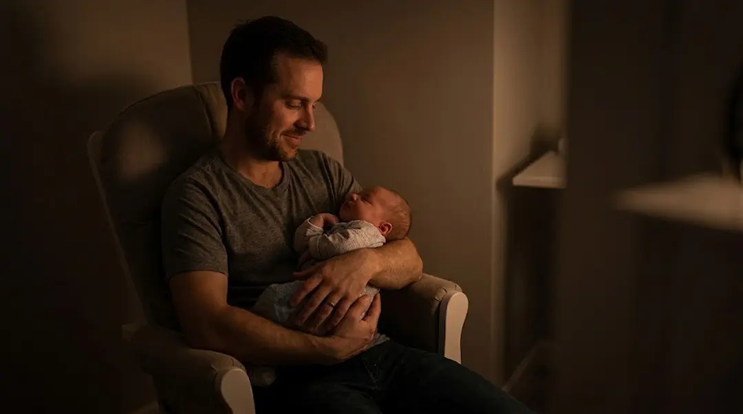 Pai sentado numa cadeira de amamentação à meia-luz, ninando o bebé adormecido nos braços durante a noite.